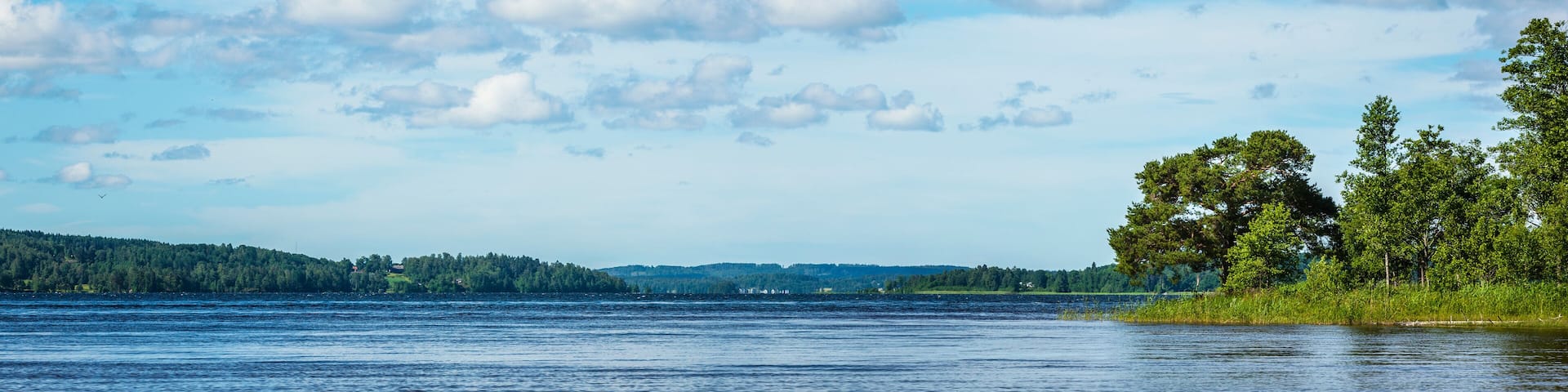 A calm lake with a forested shoreline and distant hills under a partly cloudy sky.