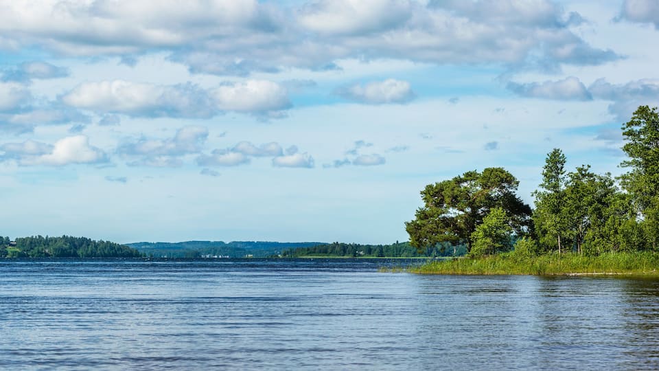 A calm lake with a forested shoreline and distant hills under a partly cloudy sky.