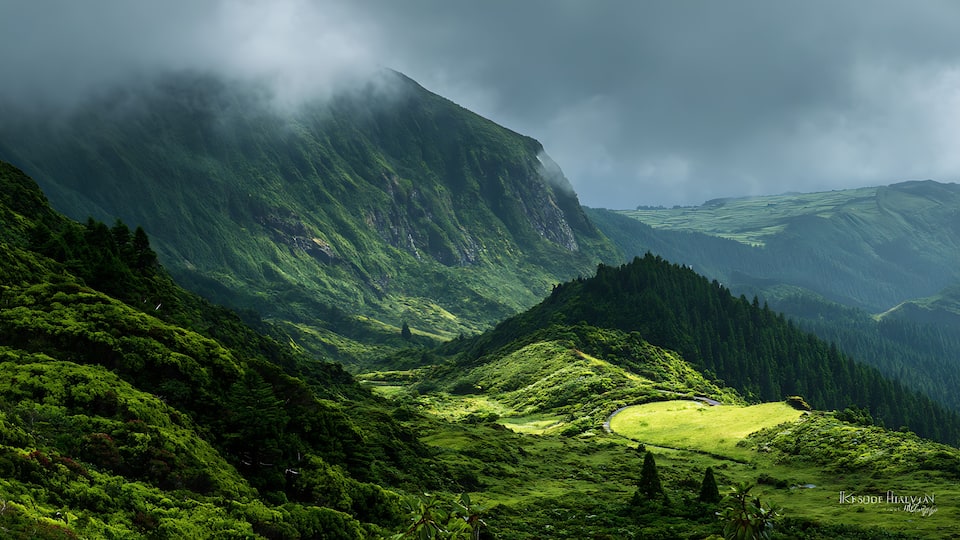 A lush green valley with a winding road, steep mountains, and a misty atmosphere.