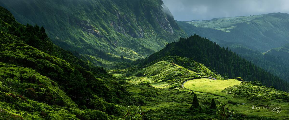 A lush green valley with a winding road, steep mountains, and a misty atmosphere.