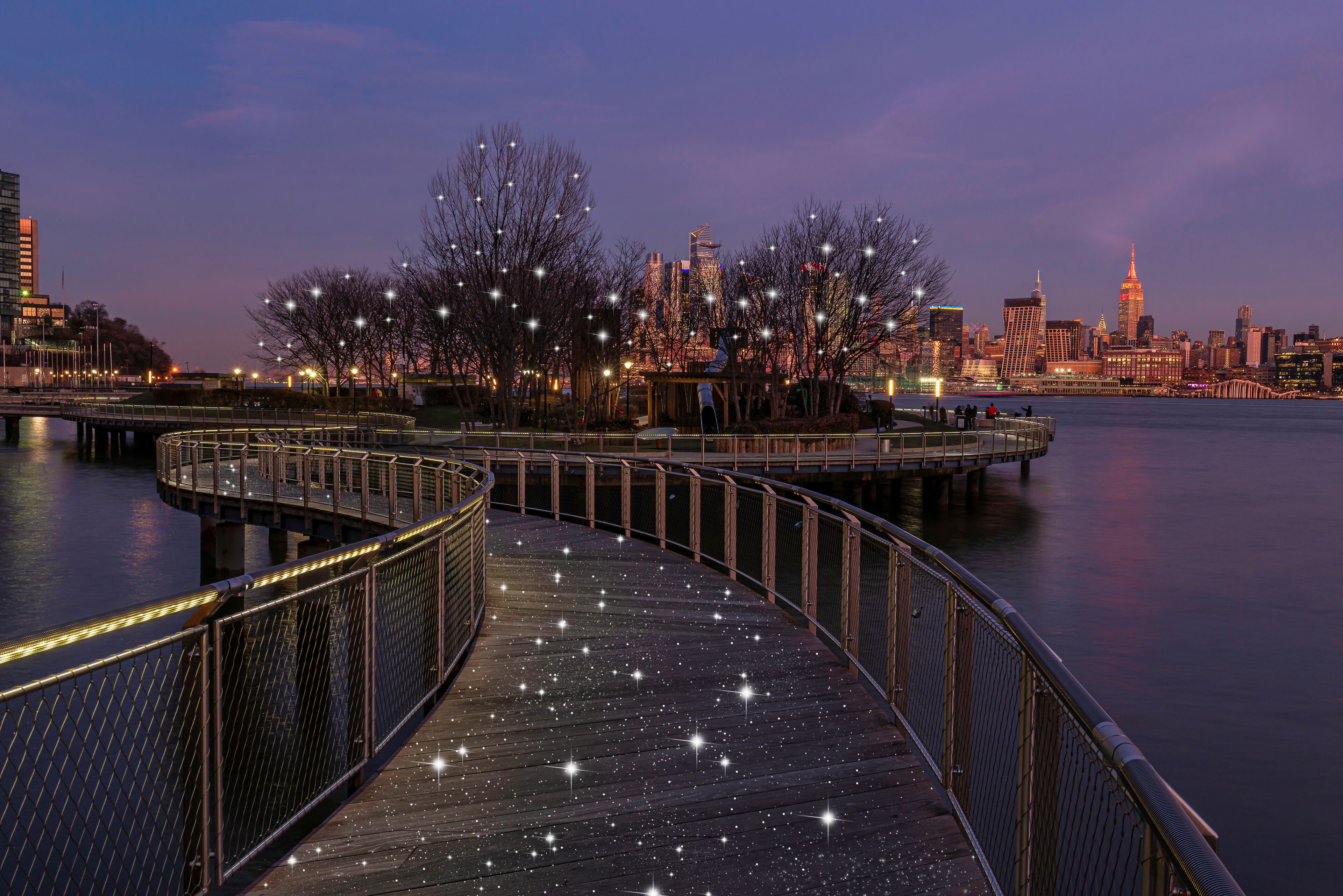A waterfront promenade with a curved walkway, city skyline, and illuminated trees against a twilight sky.