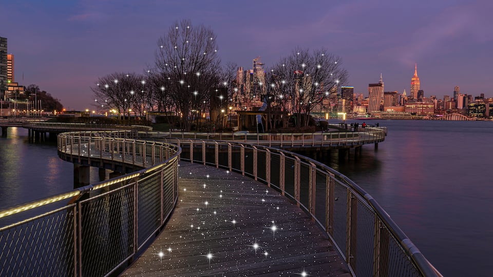 A waterfront promenade with a curved walkway, city skyline, and illuminated trees against a twilight sky.