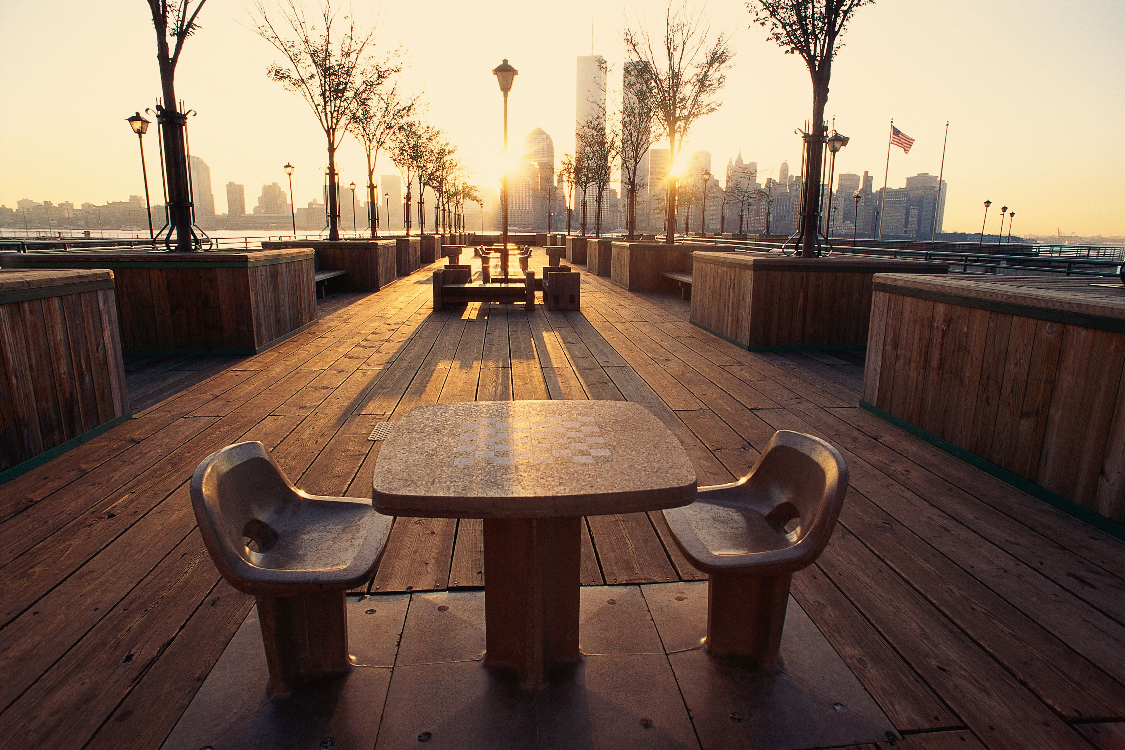A wooden boardwalk with benches and lampposts, overlooking a city skyline at sunset.
