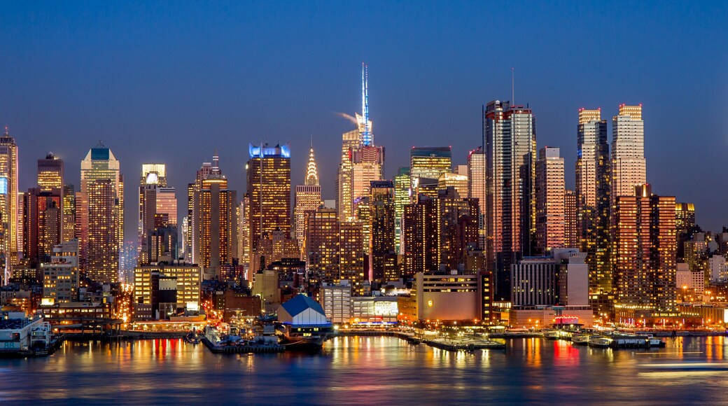 A city skyline at dusk with illuminated buildings and a river reflecting the lights.