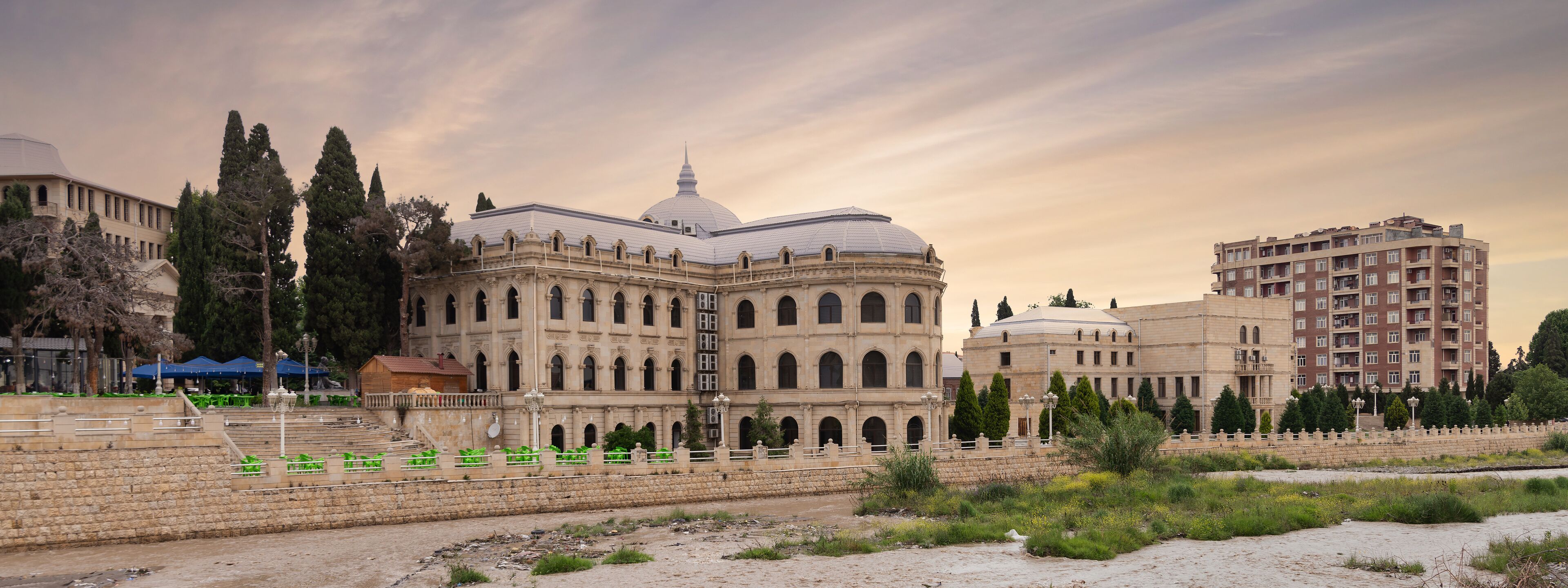 Mahsati Ganjavi Center stands majestically in Ganja, Azerbaijan, highlighted by its distinct domes against a backdrop of a dusky sky. The surrounding area features lush greenery and modern buildings