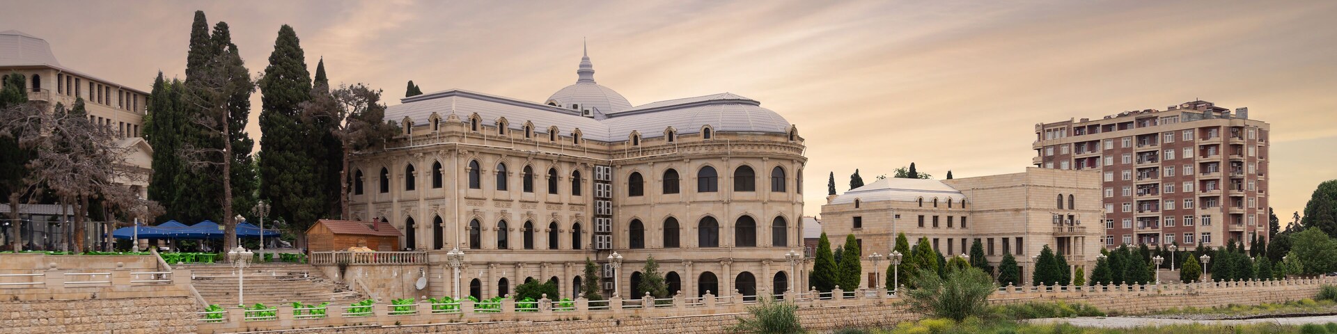 Un edificio histórico con cúpula y ventanas arqueadas, rodeado de árboles y un río.