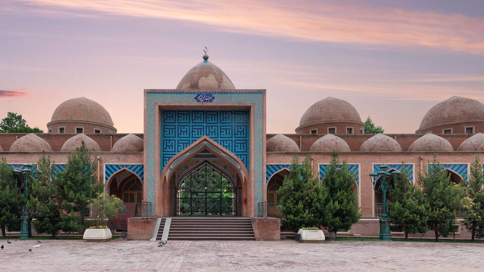 A mosque with domed architecture, a central entrance with a blue door, and a surrounding courtyard with trees and pigeons.