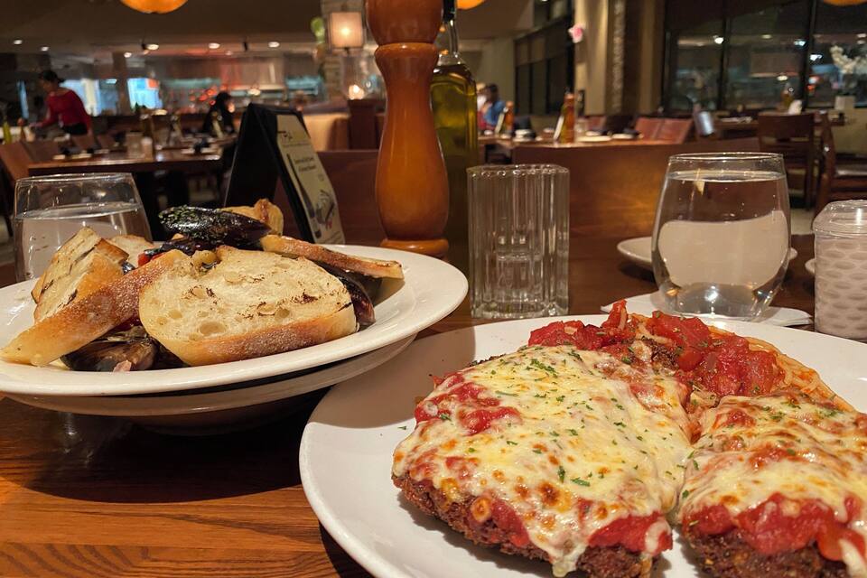A restaurant setting with a plate of bread and mushrooms, a plate of pizza, and a bottle of olive oil on the table.