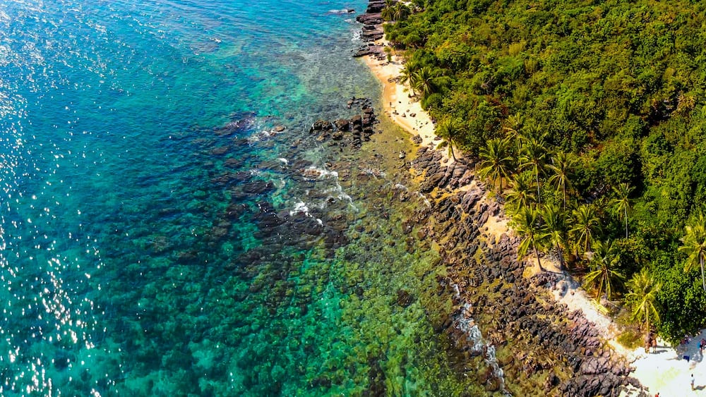 A coastal landscape with clear turquoise waters, a sandy beach, and dense tropical vegetation.