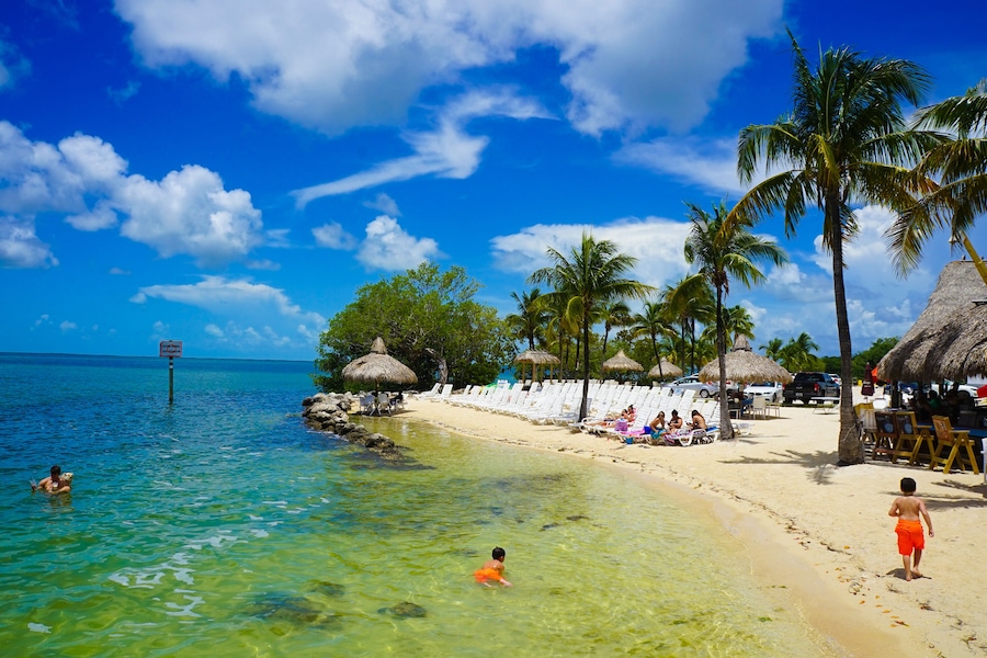 Une plage à l’eau turquoise et limpide, bordée de palmiers et ombragée par des parasols en feuilles de palmier.
