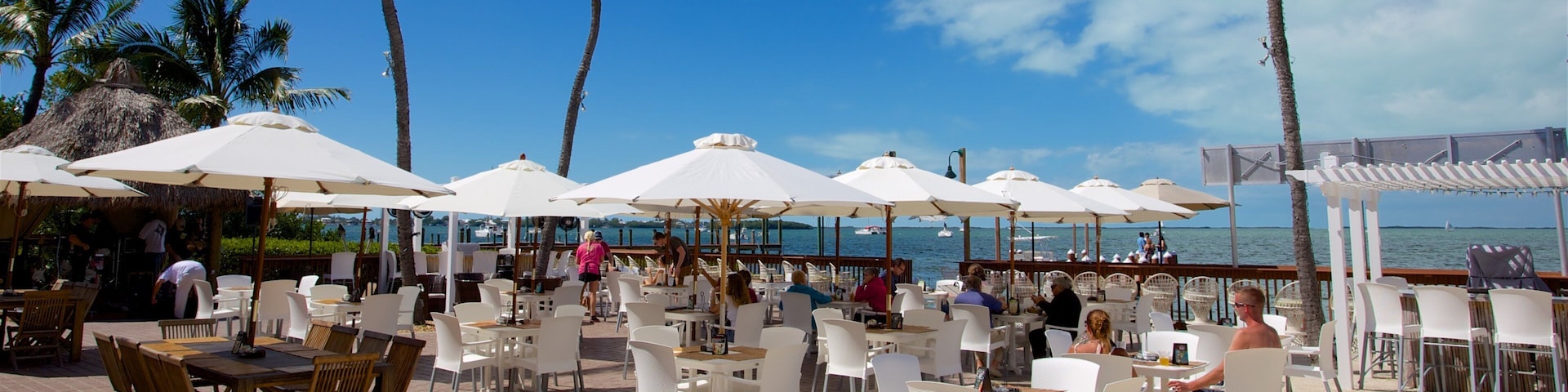 A beachside restaurant with white tables and chairs, palm trees, and a view of the sea.