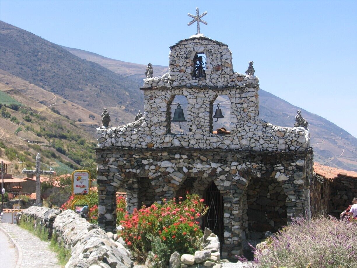 A stone chapel with a bell tower, surrounded by a stone wall and flowering plants.