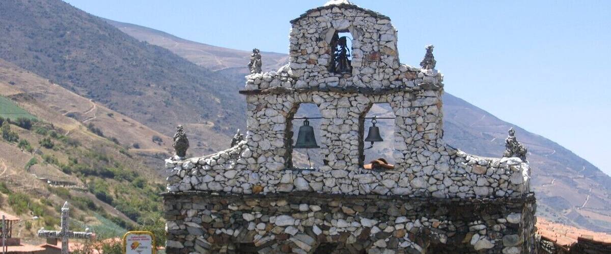 A stone chapel with a bell tower, surrounded by a stone wall and flowering plants.