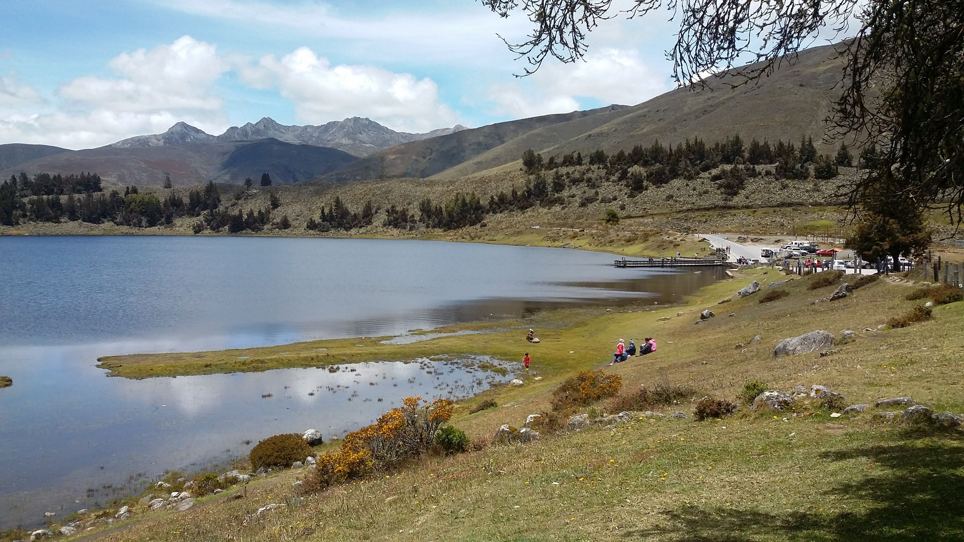 A lake surrounded by mountains, a dock, and people near the shore.