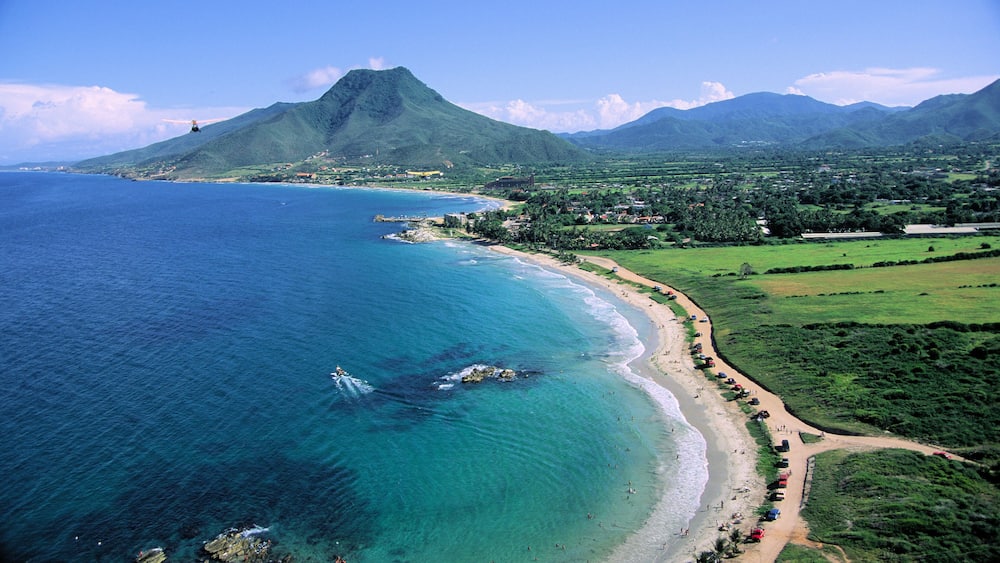 A coastal landscape with a beach, clear blue waters, and a mountain in the background.