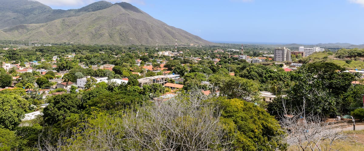 A coastal town with mountains in the background, surrounded by dense greenery and red-roofed buildings.