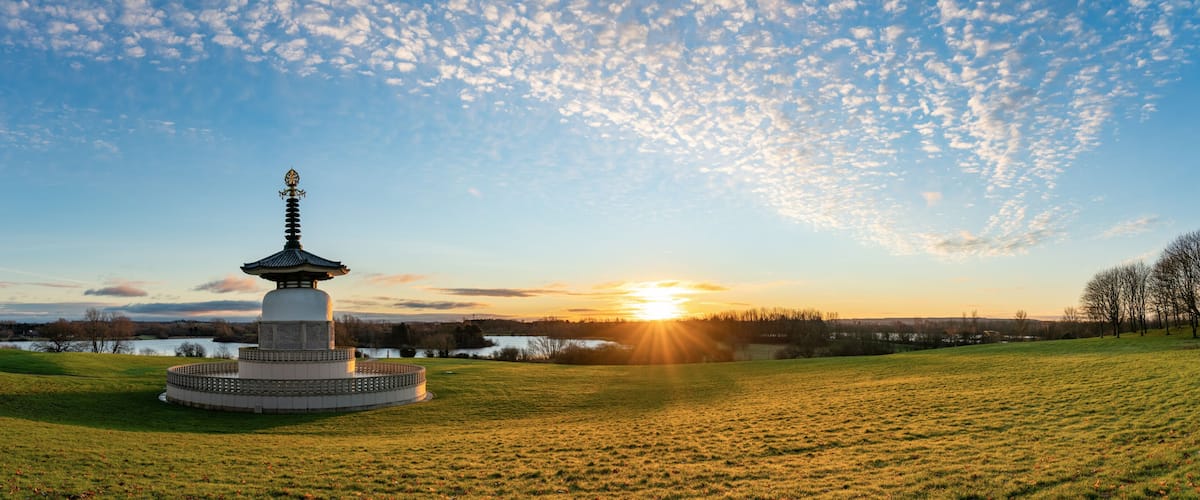 A pagoda-style monument in a grassy field with a lake and sunset in the background.