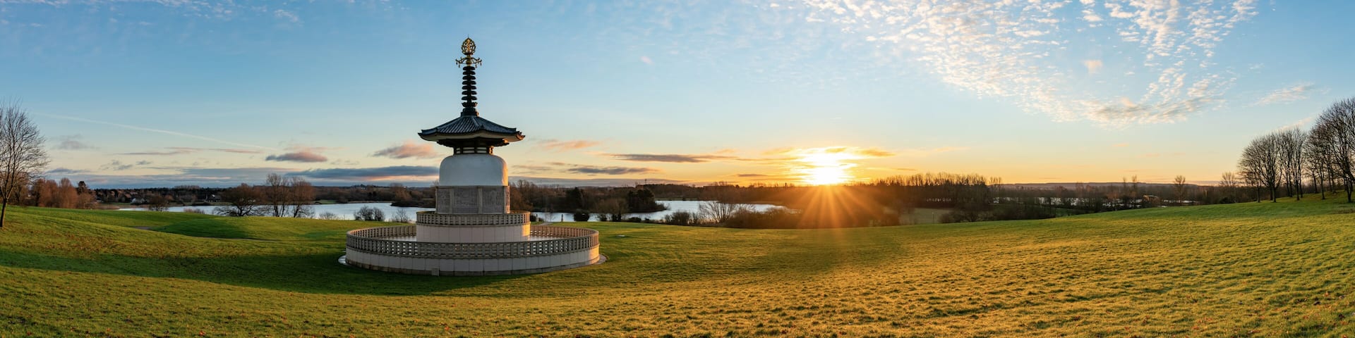 A pagoda-style monument in a grassy field with a lake and sunset in the background.