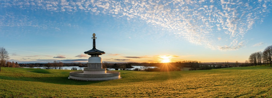 Un monumento de estilo pagoda en un campo cubierto de hierba, con un lago y el atardecer al fondo.