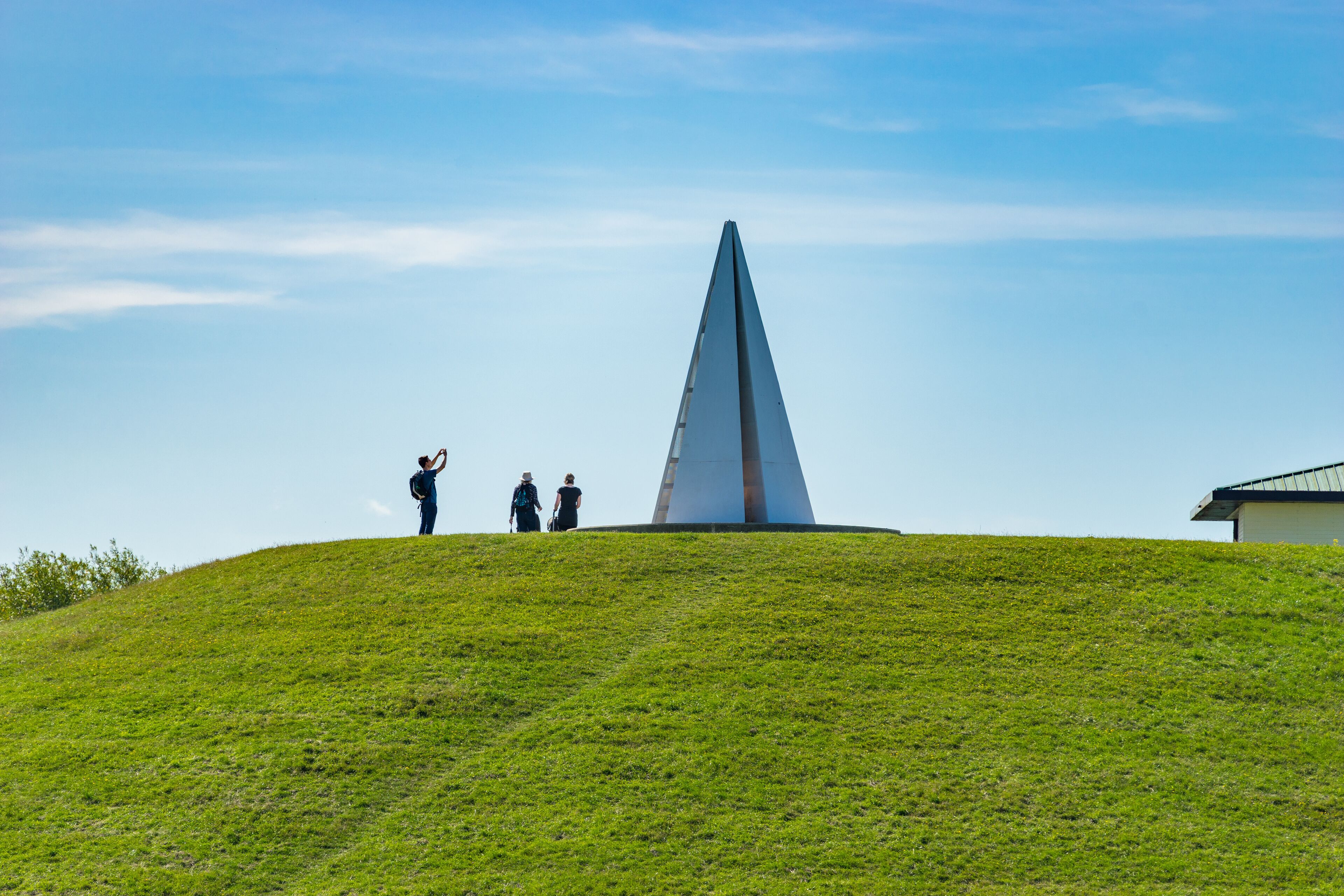 A grassy hill with a tall, triangular monument and a small building in the background.