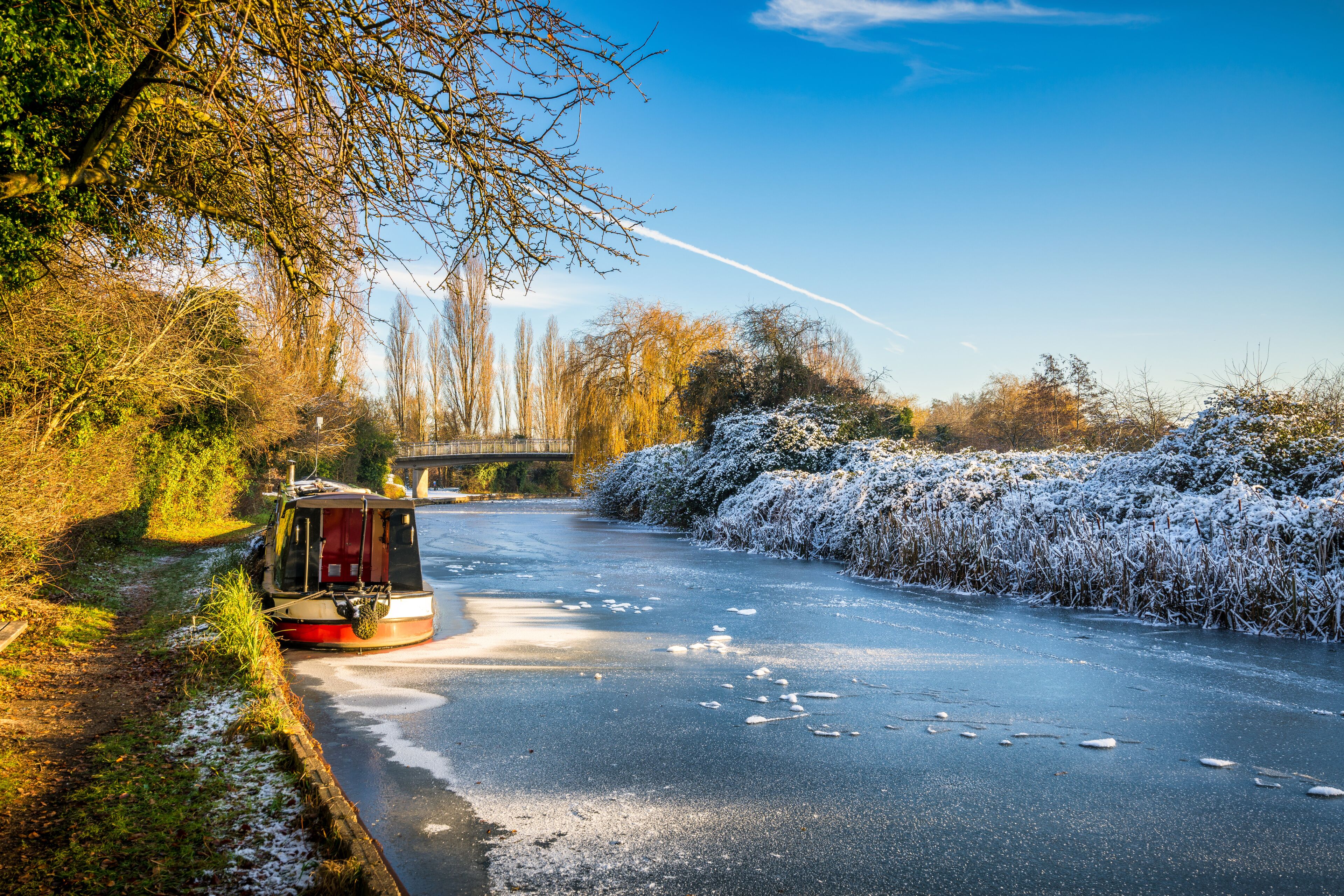 A canal with a boat, snow-covered bushes, and a bridge in the background.