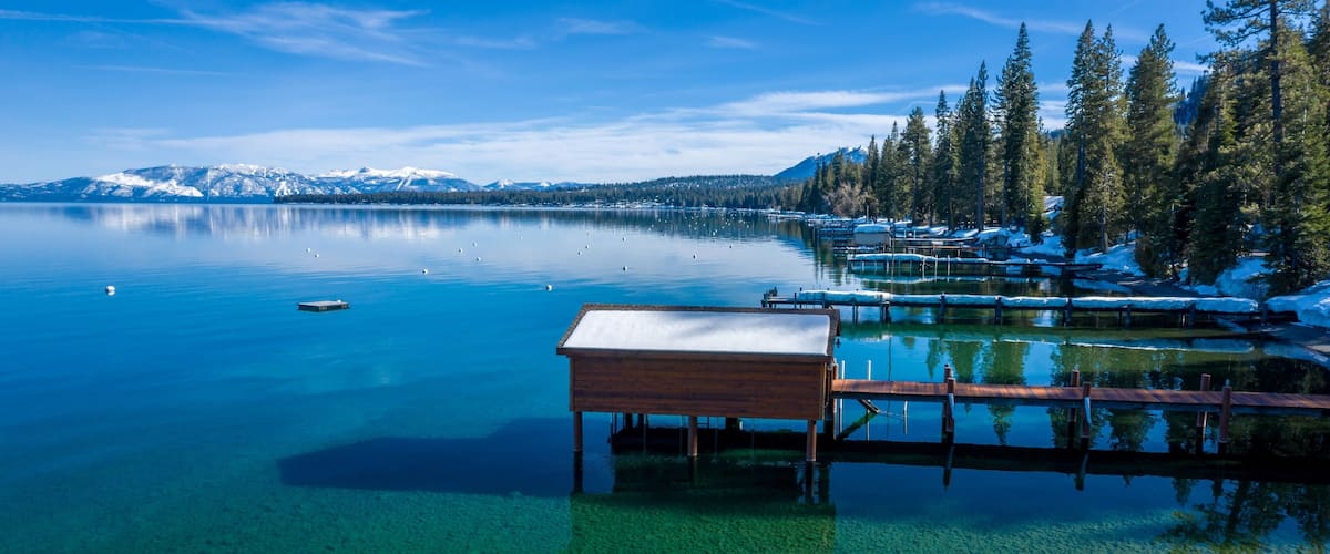 A clear lake with a wooden dock, snow-covered mountains, and evergreen trees in the background.