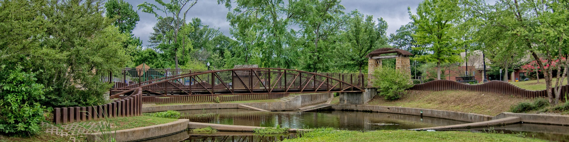 Un parque con un puente metálico sobre un estanque, rodeado de árboles y un edificio de piedra.
