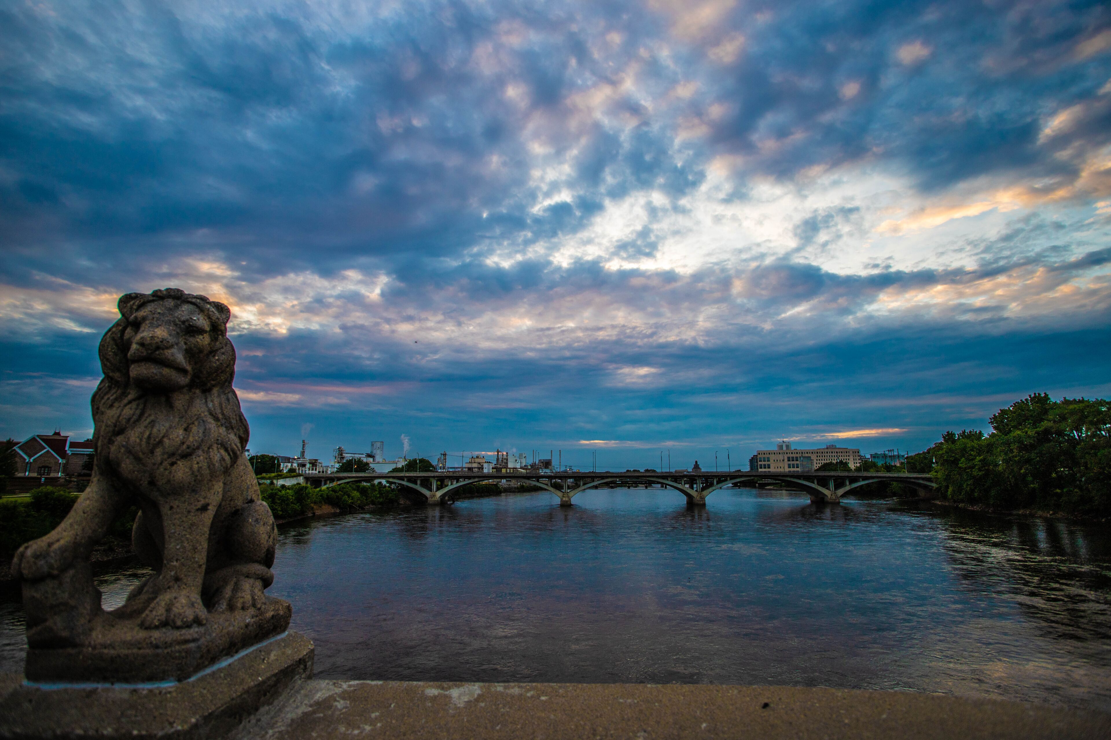 A stone lion statue by a river with a bridge in the background.