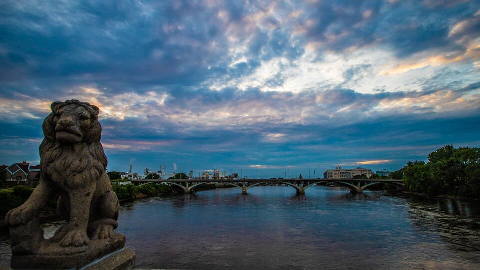 A stone lion statue by a river with a bridge in the background.