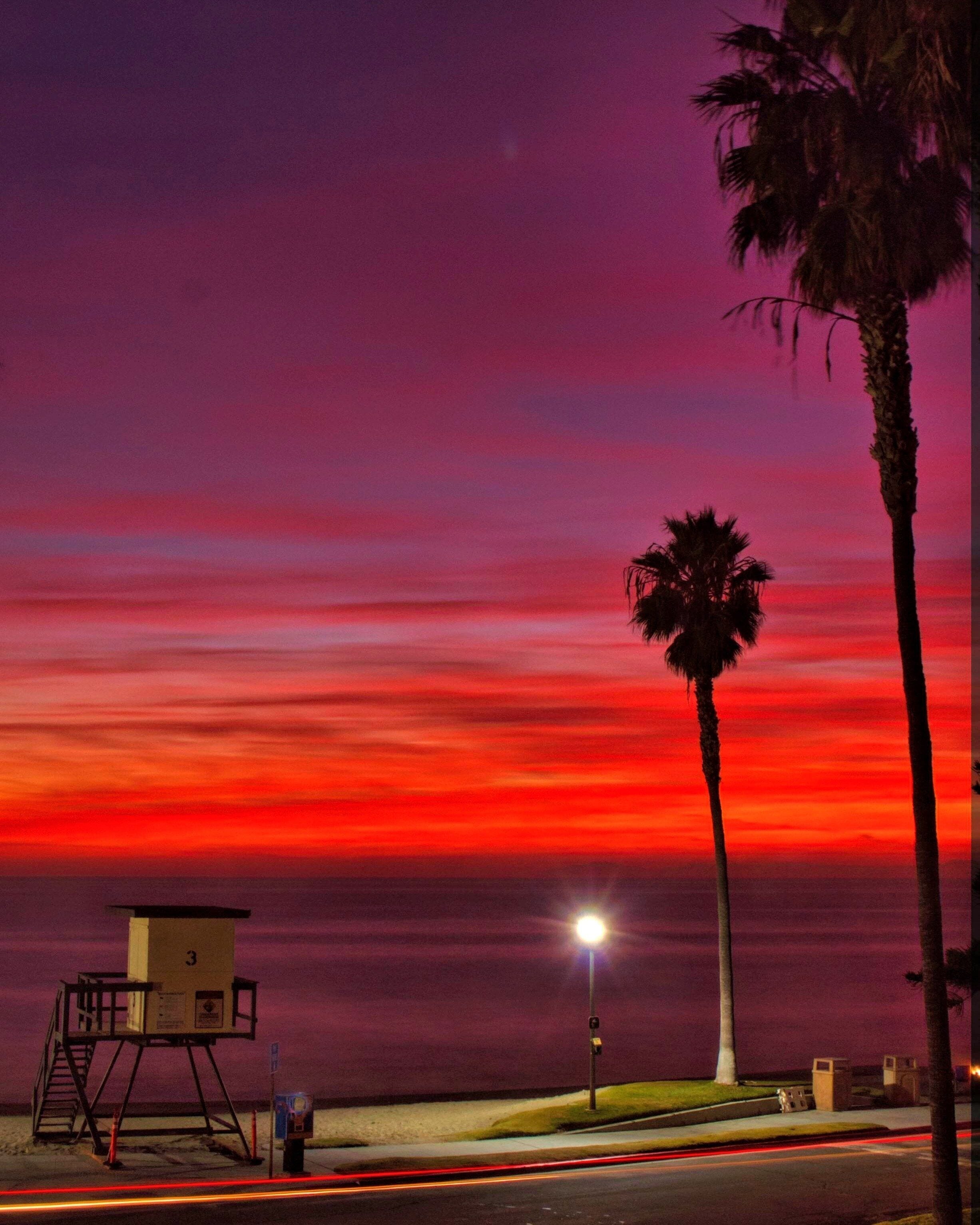 A sunset with a red sky, palm trees, a lifeguard tower, and a street with light trails.