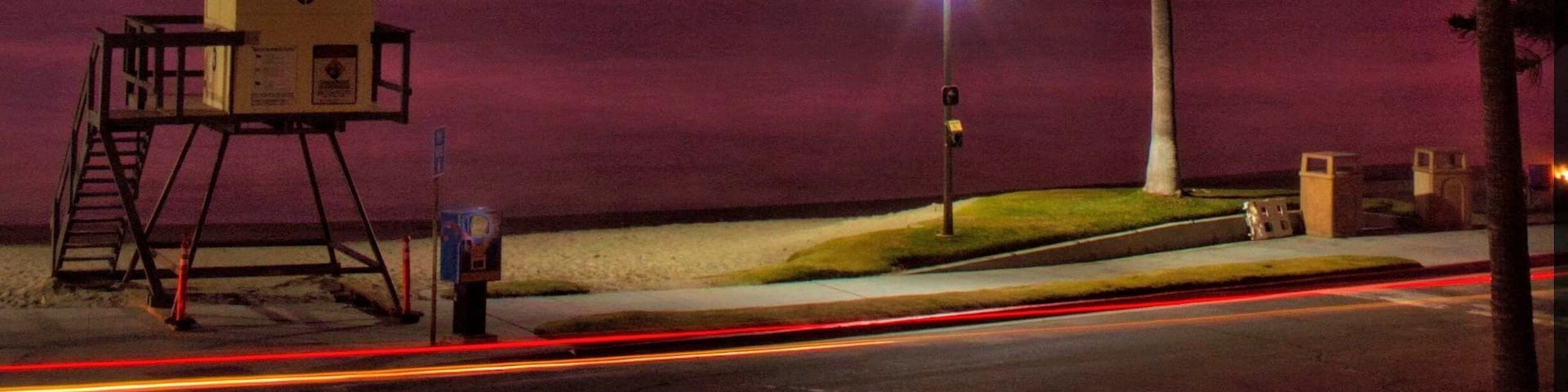 Un coucher de soleil avec un ciel rouge, des palmiers, une tour de surveillance des sauveteurs et une rue illuminée par des traînées de lumière.