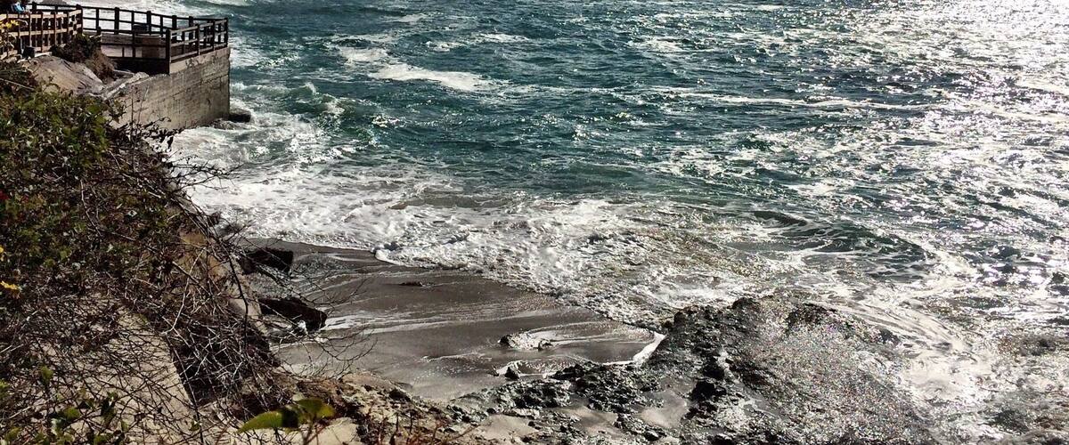 A coastal landscape with waves crashing onto the shore, a wooden walkway, and a mountainous backdrop.