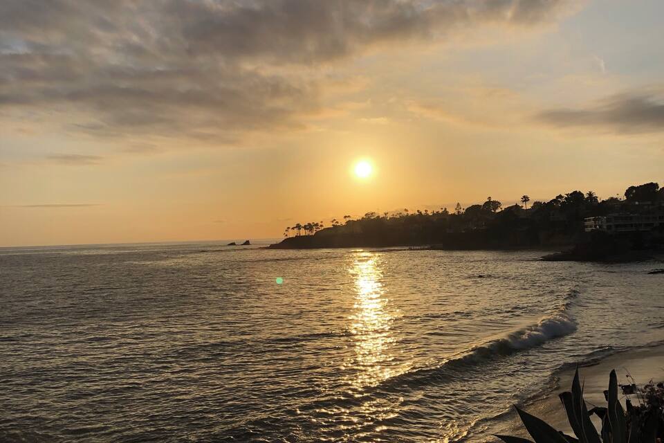 A sunset over a coastal area with a beach, ocean, and a distant shoreline with buildings and palm trees.