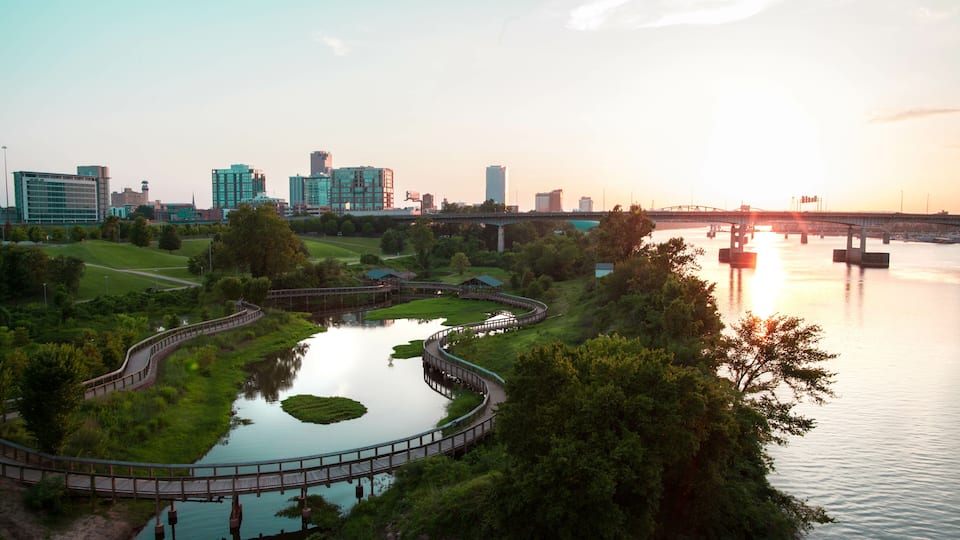 A riverside park with a curved walking path, a bridge, and a city skyline in the background.