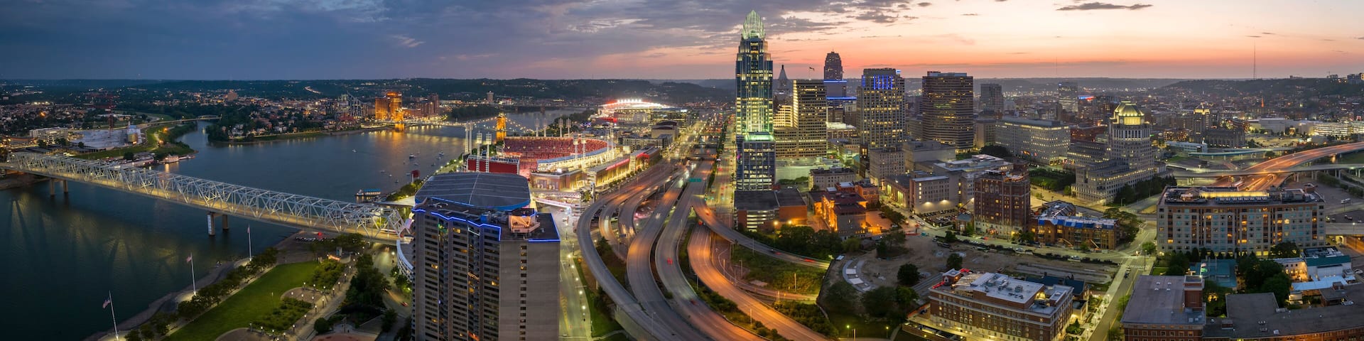 A cityscape with a river, a bridge, and a stadium at dusk.