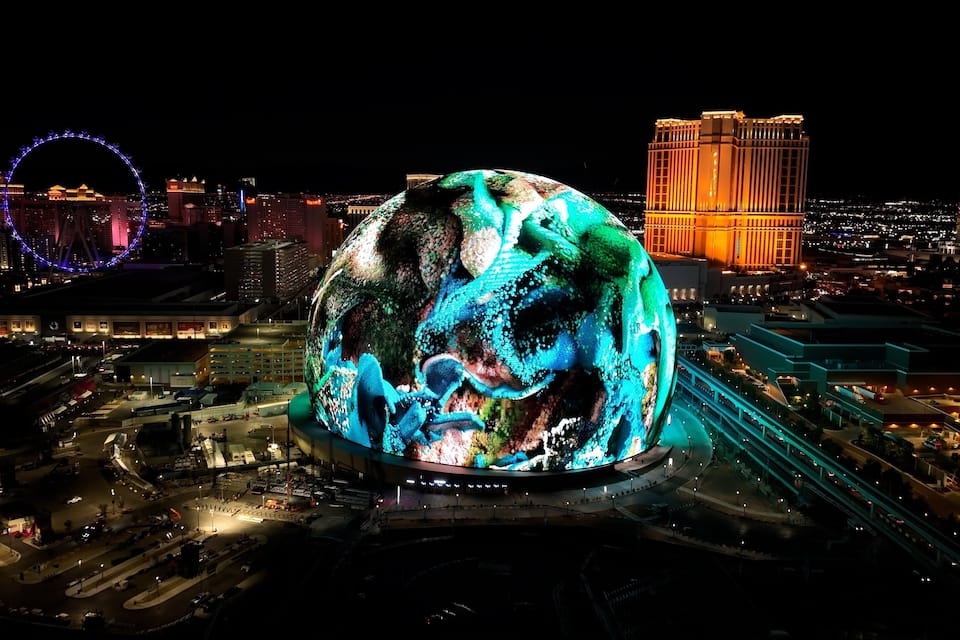 A large illuminated globe sculpture in an urban setting with surrounding buildings and a Ferris wheel in the background.