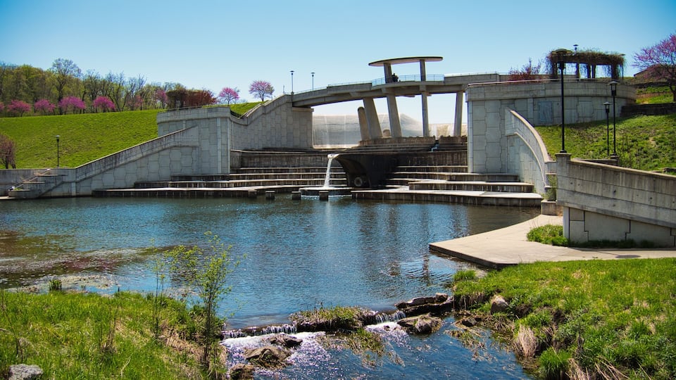 A stone bridge over a calm river with a small waterfall, surrounded by green grass and trees.
