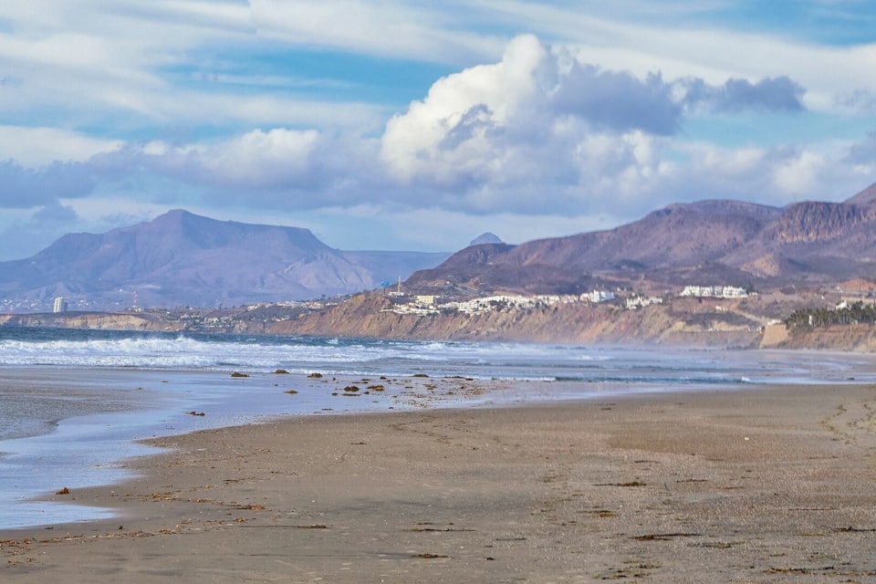 A beach with waves, mountains, and coastal buildings.