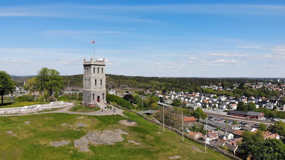 A historic tower with a flag, surrounded by a park with a stone wall, in a town with houses and a railway.