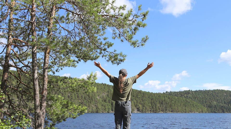 A person standing by a lake with trees and a forest in the background.