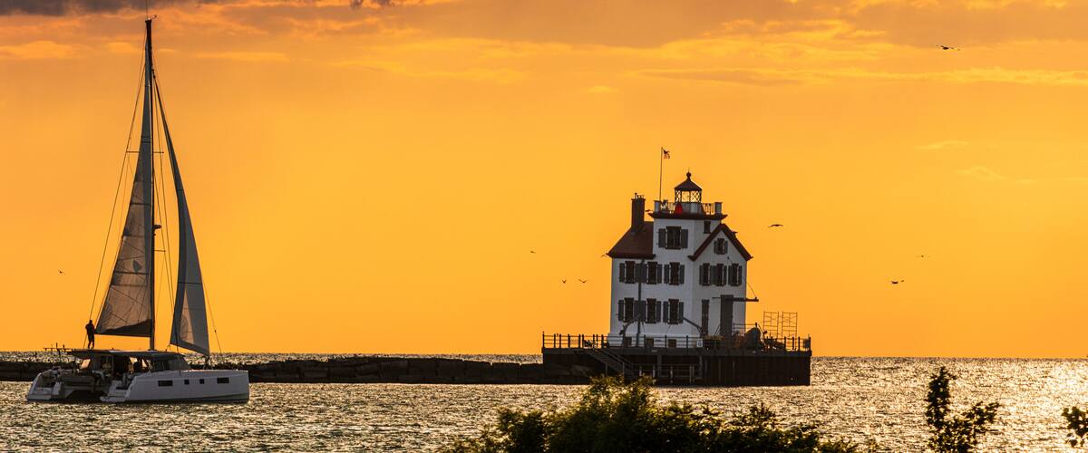 A sailboat on the water with a lighthouse in the background during sunset.