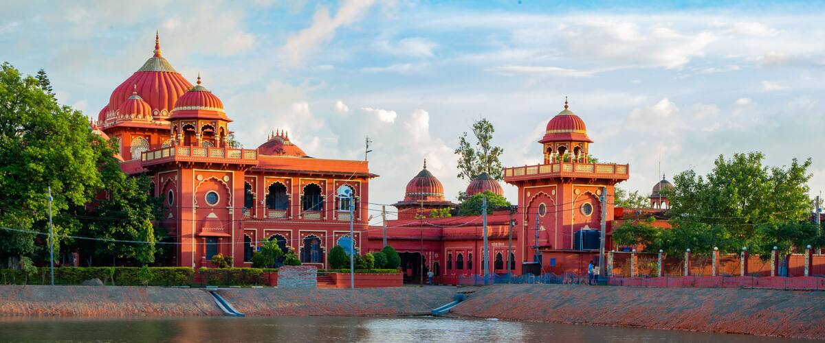 A large, ornate building with multiple domes and arches, situated beside a calm body of water.