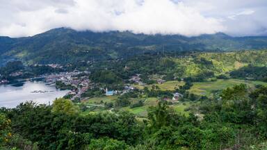 Paisagem montanhosa com uma cidade à beira de um lago, rodeada por vegetação exuberante e colinas suaves.