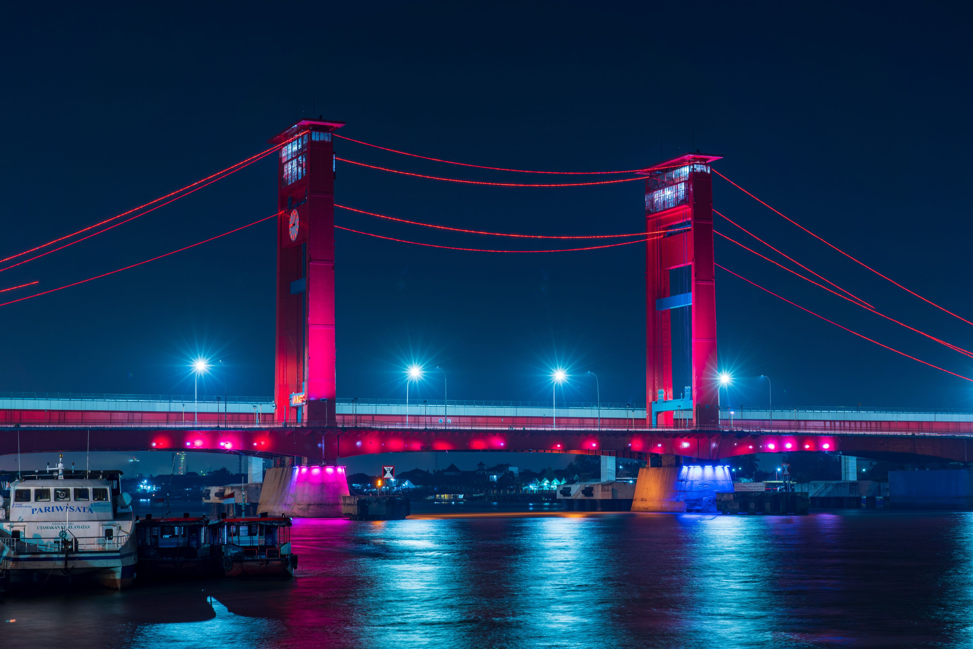 Un pont ben il·luminat, amb cables i llums vermelles, un riu a sota i embarcacions amarrades a la riba.