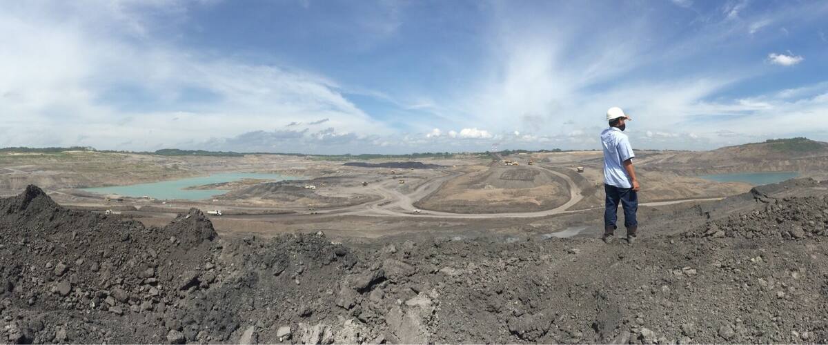 A person standing on a rocky terrain overlooking a mining area with a large open pit and a body of water.