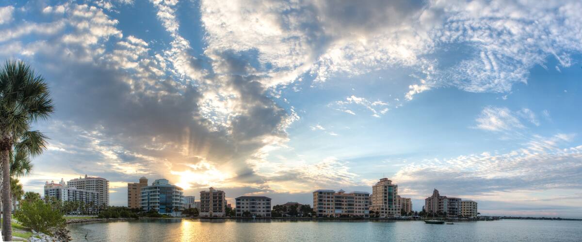 A coastal cityscape with a sunset over the water, featuring several high-rise buildings and palm trees.