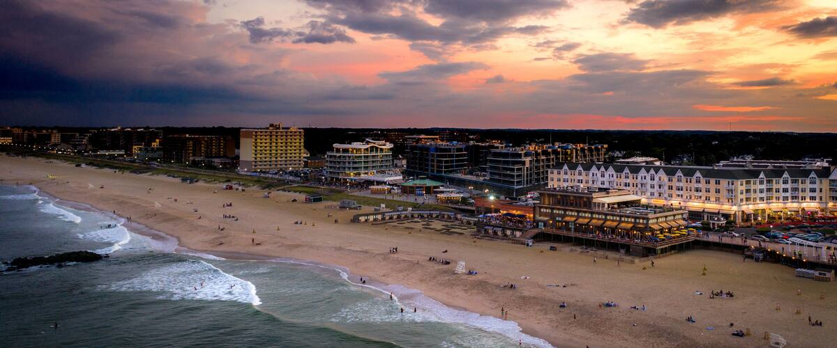 Een kuststreeaatbeeld met een strand, oceaan golven en diverse gebouwen, waaronder hotels en restaurants.