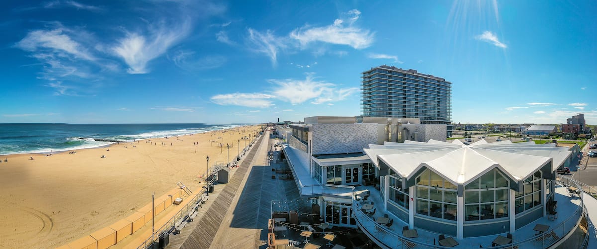 A beach with a boardwalk, a modern building with a unique roof, and a tall hotel in the background.