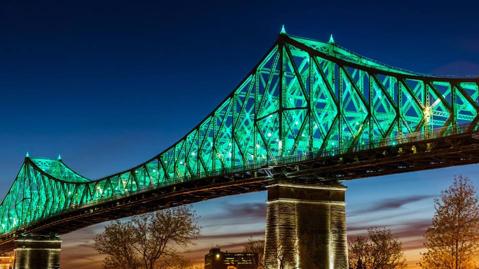 A large bridge with green lighting, spanning over a body of water with city buildings in the background.