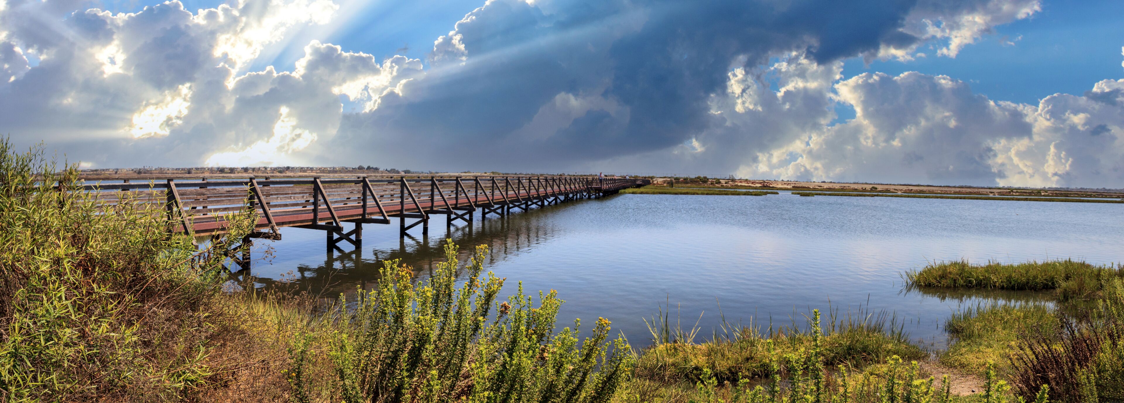A wooden bridge over a calm body of water, surrounded by greenery and under a cloudy sky.